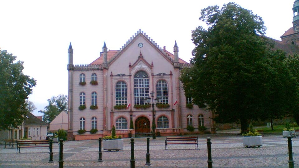 A pink building with arched windows and a clock, surrounded by cobblestone pavement and benches.