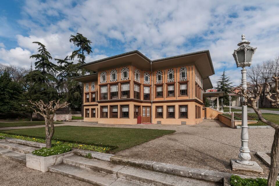 Un grand immeuble à étages, doté d’une façade beige et de fenêtres richement décorées, entouré d’un jardin bien entretenu et d’arbres.