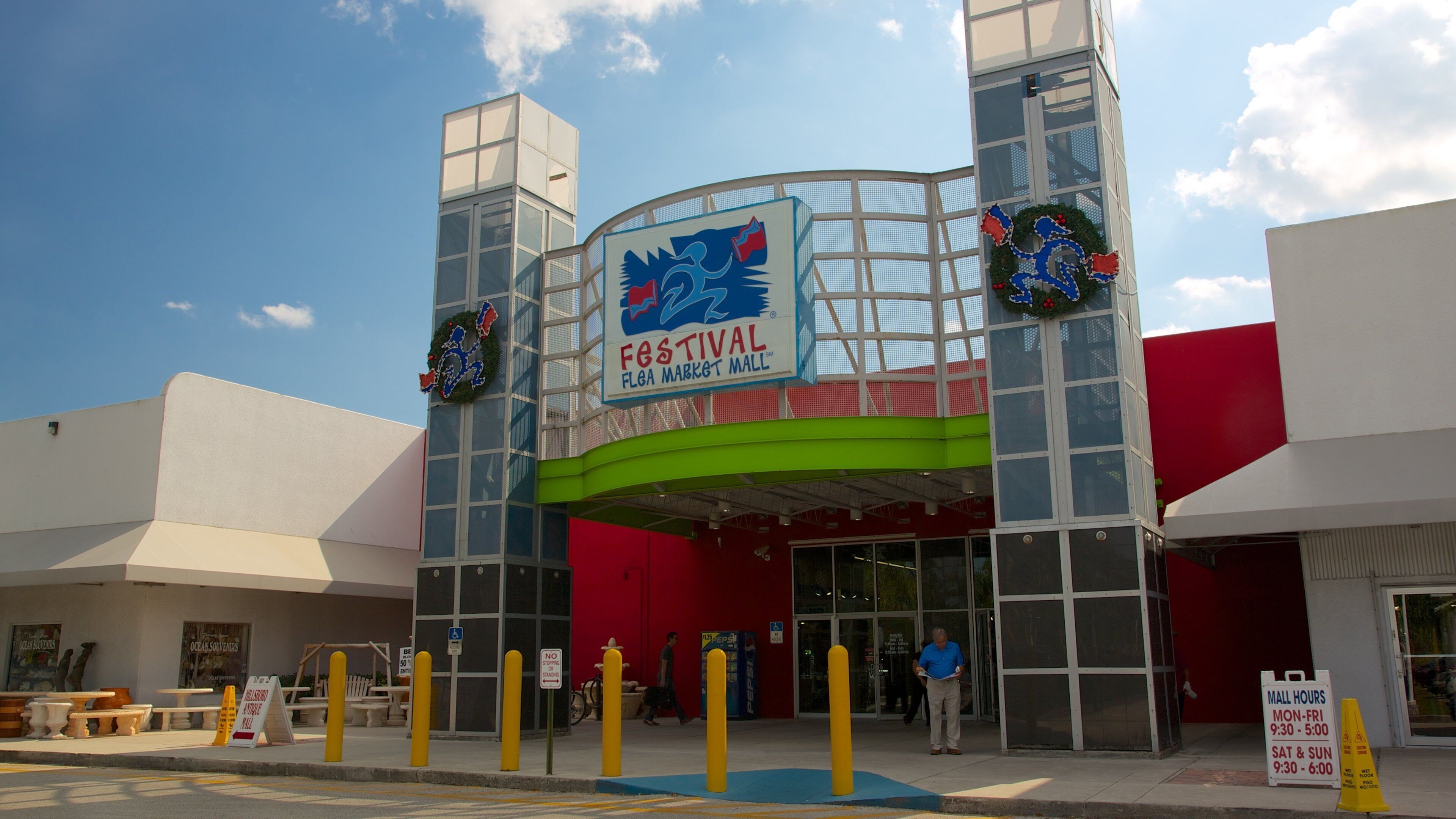 The exterior of Festival Flega Market Mall with a large sign, two decorative wreaths, and a blue ramp leading to the entrance.
