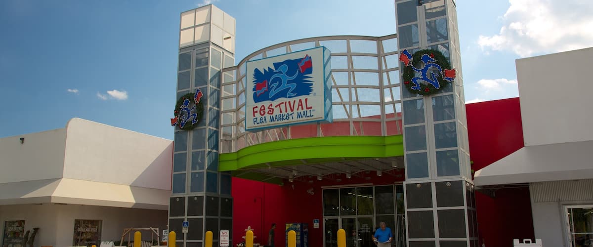 The exterior of Festival Flega Market Mall with a large sign, two decorative wreaths, and a blue ramp leading to the entrance.