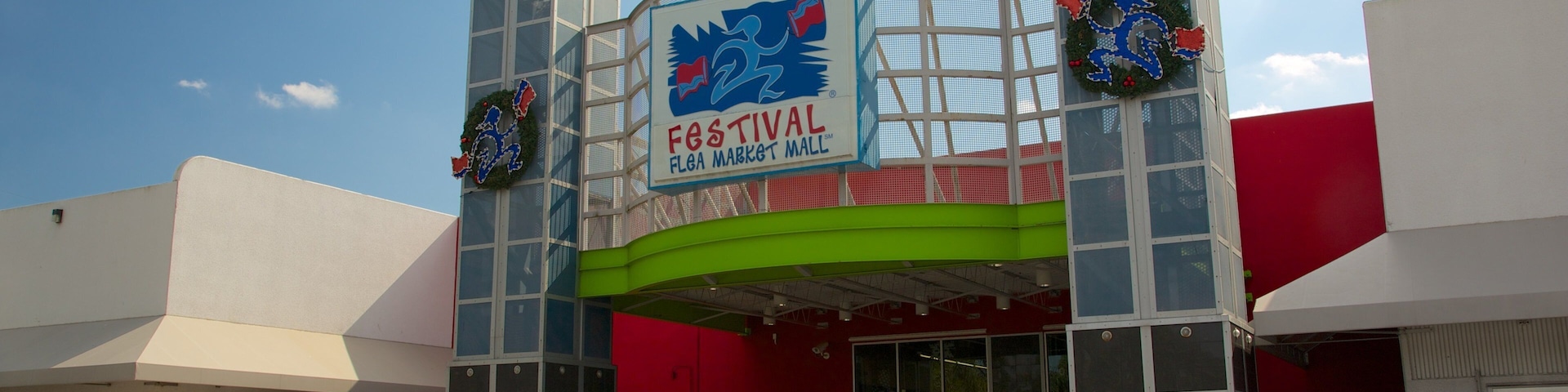 The exterior of Festival Flega Market Mall with a large sign, two decorative wreaths, and a blue ramp leading to the entrance.