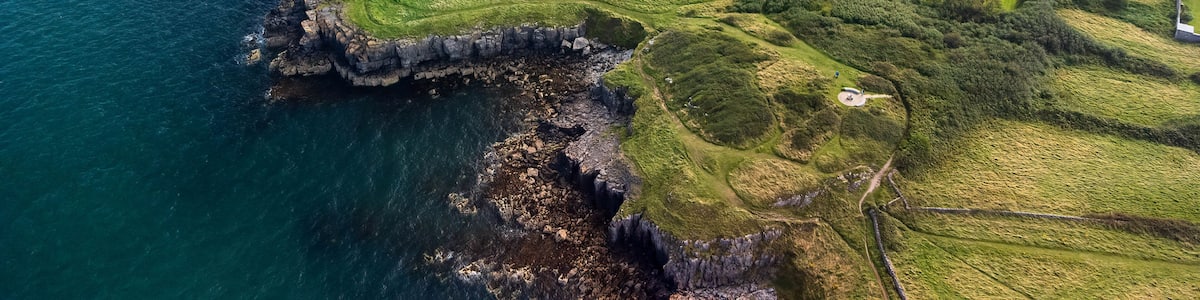 Eine Luftaufnahme einer Küstenlandschaft mit steilen Felsen, grünen Wiesen und einem kleinen Haus am Wasser.