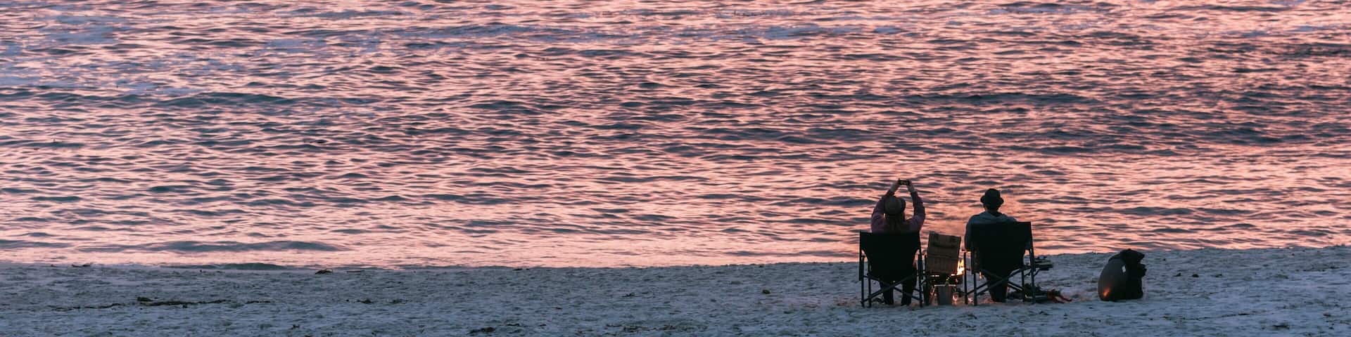 A beach with people sitting on chairs facing the ocean during sunset.