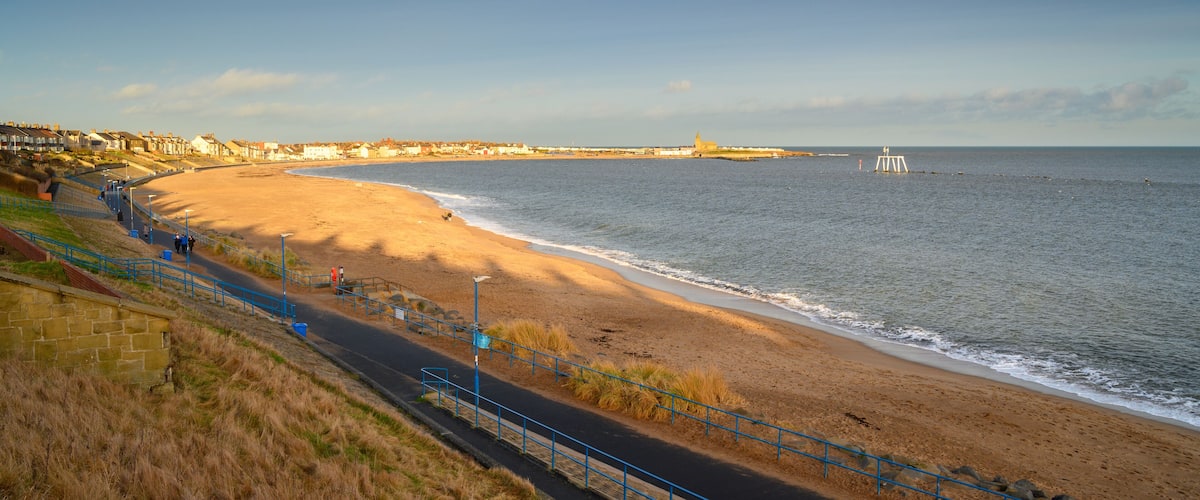 A coastal road with a beach, a lighthouse, and residential buildings.