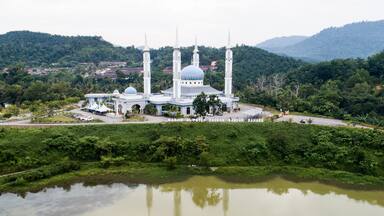 Uma mesquita com uma grande cúpula central e quatro minaretes, cercada por vegetação exuberante e um corpo de água.