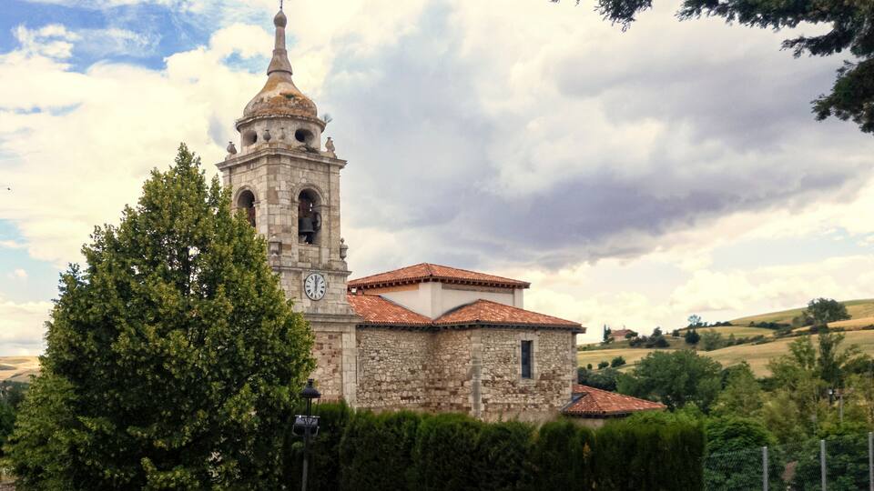 A stone church with a bell tower and a clock, surrounded by trees and hills.