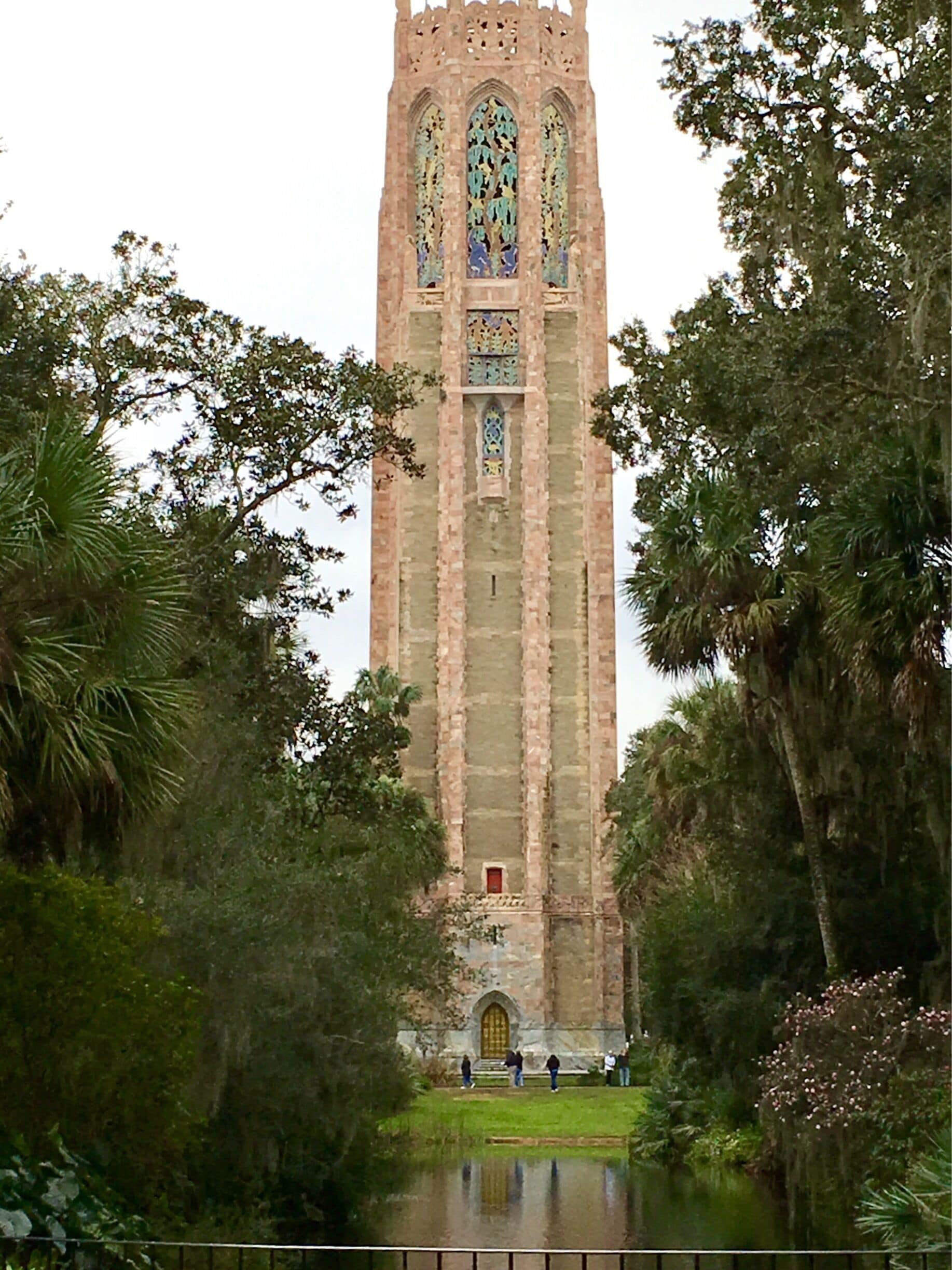 A tall, Gothic-style tower with intricate stained glass windows, surrounded by lush greenery and a small pond.