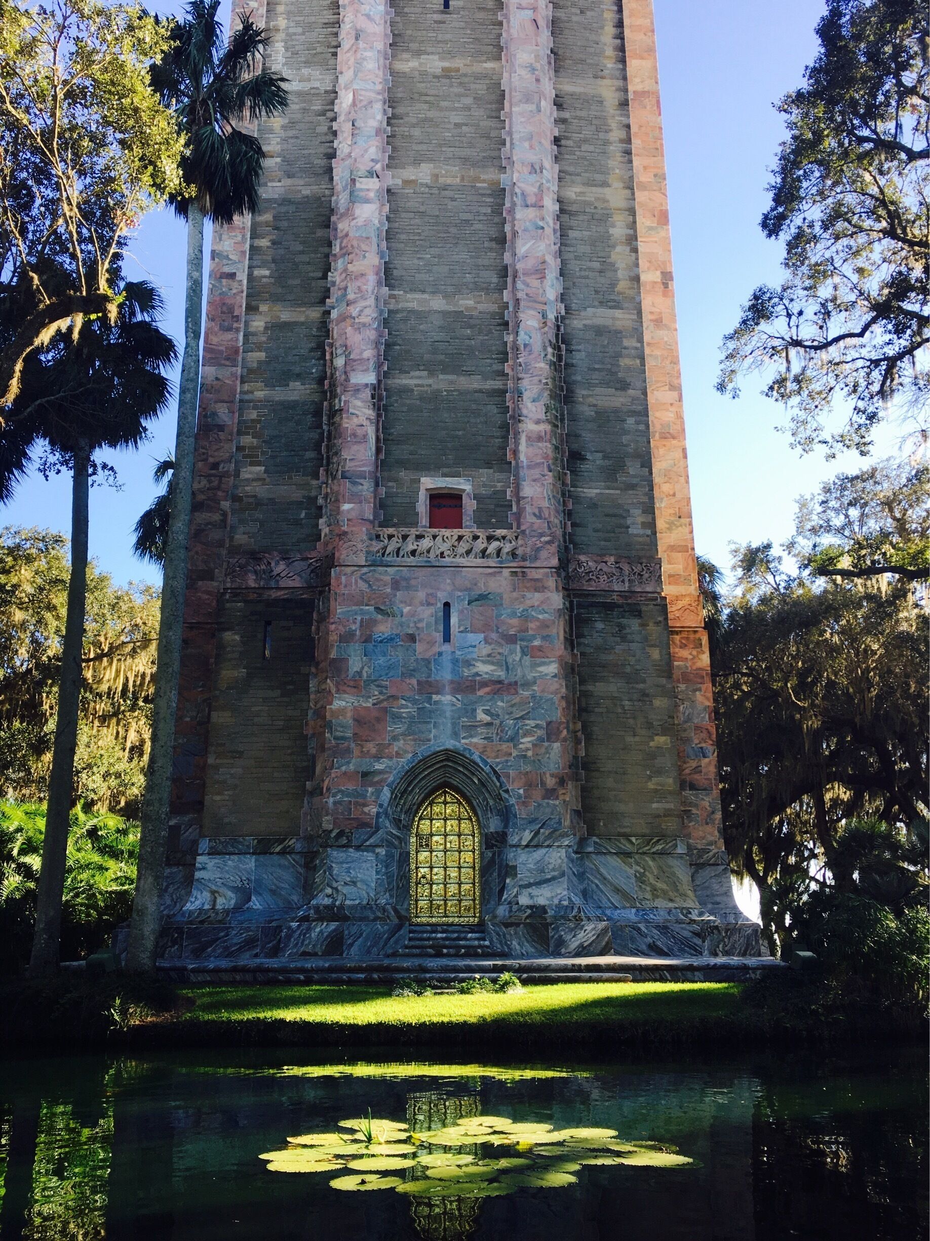 A tall stone tower with a golden door and a small window, surrounded by trees and a pond.