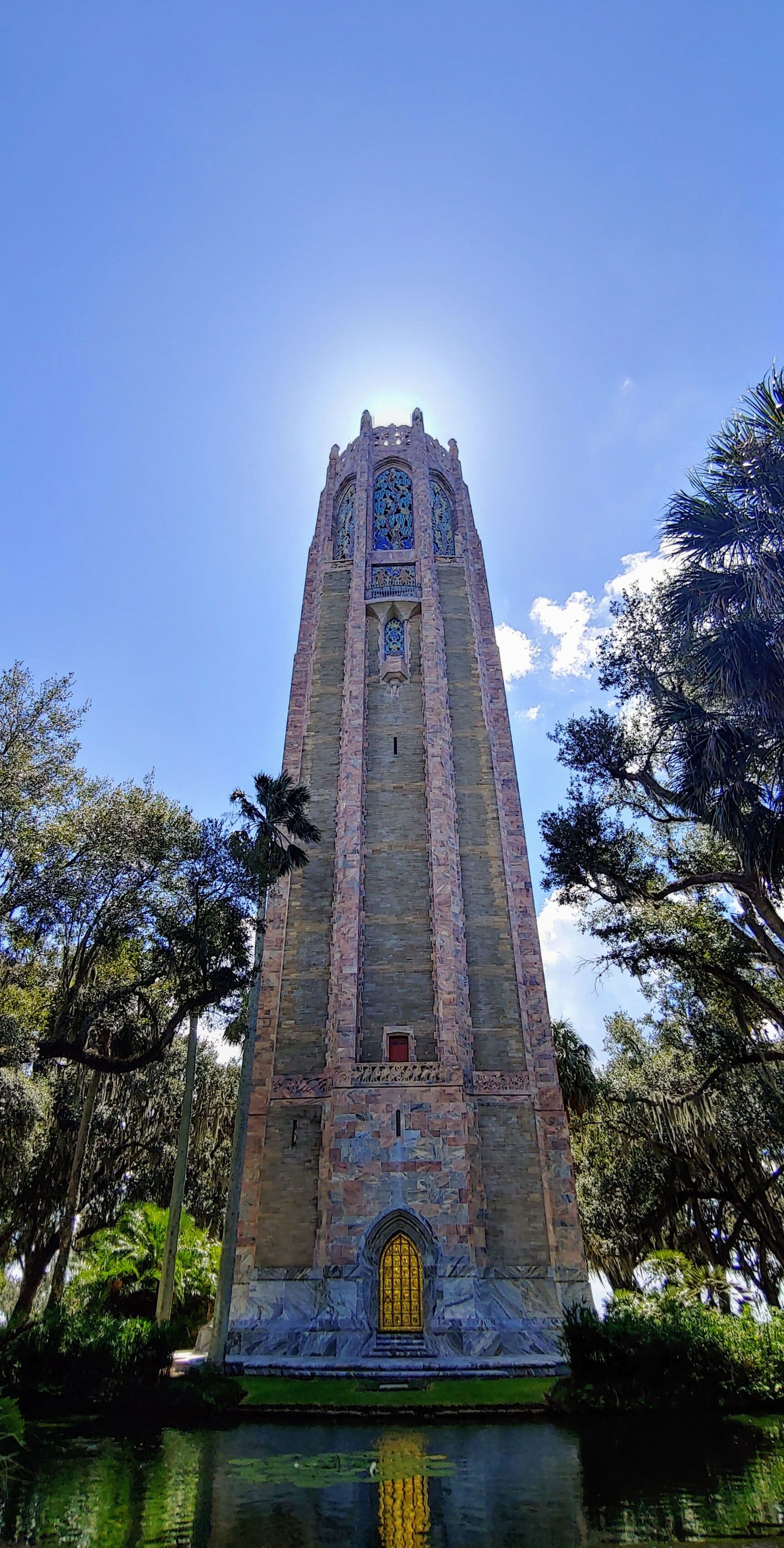 A tall stone tower with intricate stained glass windows and a golden door, surrounded by lush greenery and a reflective water body.