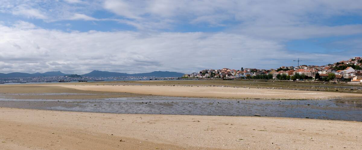 A coastal beach with a sandy shore, a cityscape in the background, and mountains in the distance.