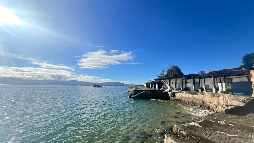A waterfront with a pier extending into calm waters, a clear sky, and distant mountains.