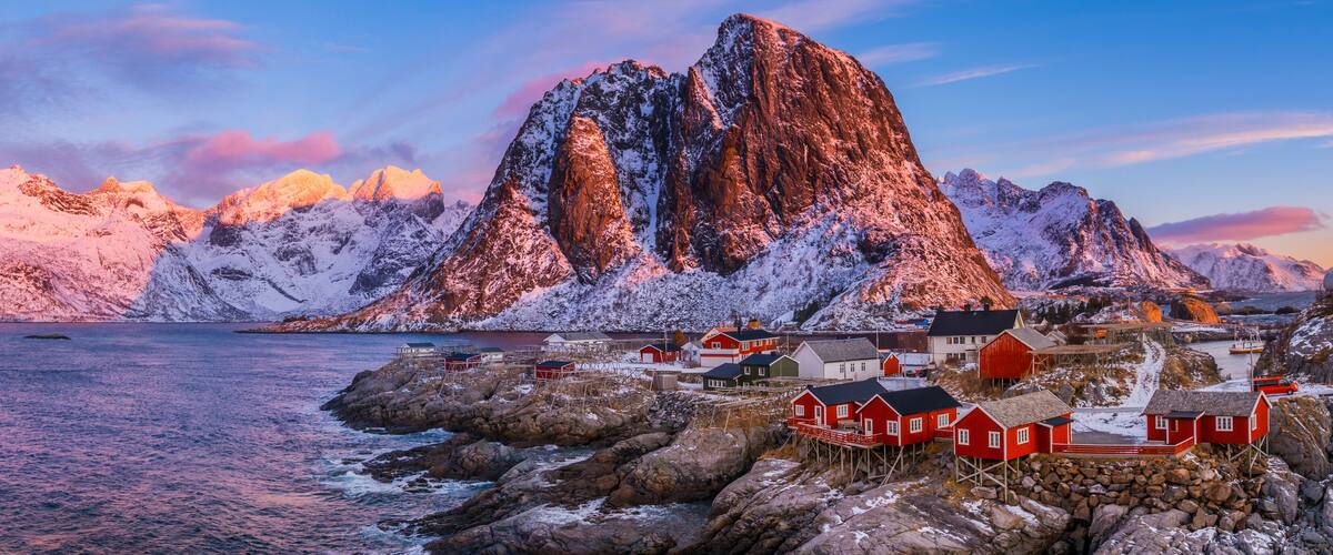 A coastal village with red houses on stilts, surrounded by snow-covered mountains and a calm sea.