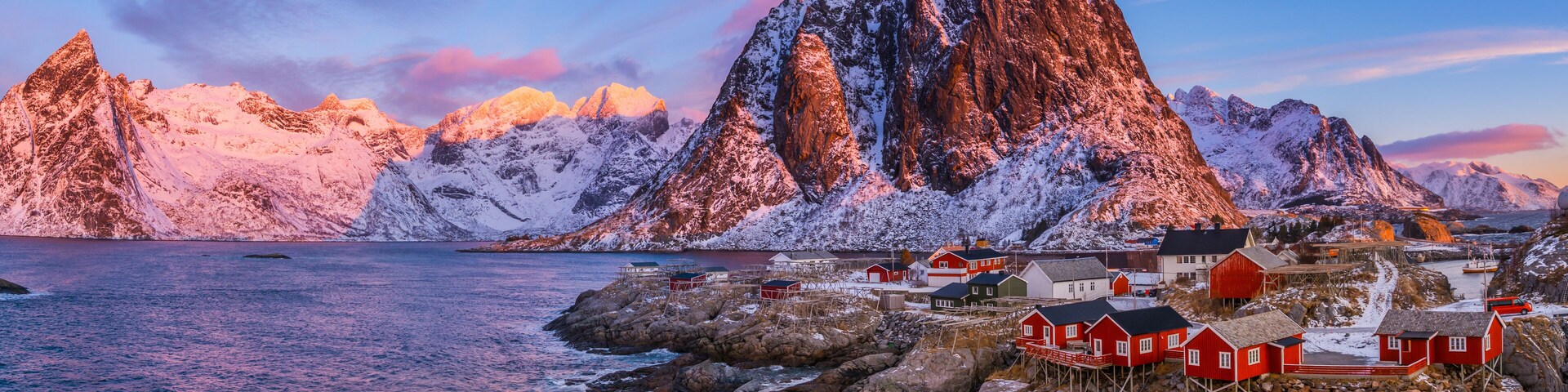 A coastal village with red houses on stilts, surrounded by snow-covered mountains and a calm sea.