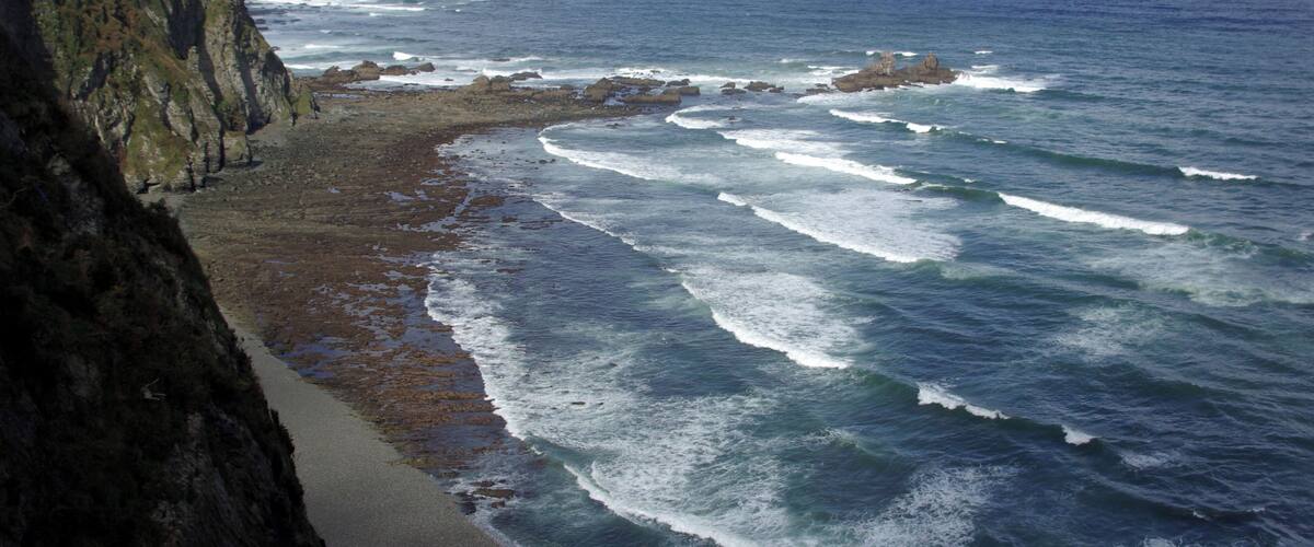 A coastal landscape with a rocky cliff, a sandy beach, and waves crashing onto the shore.