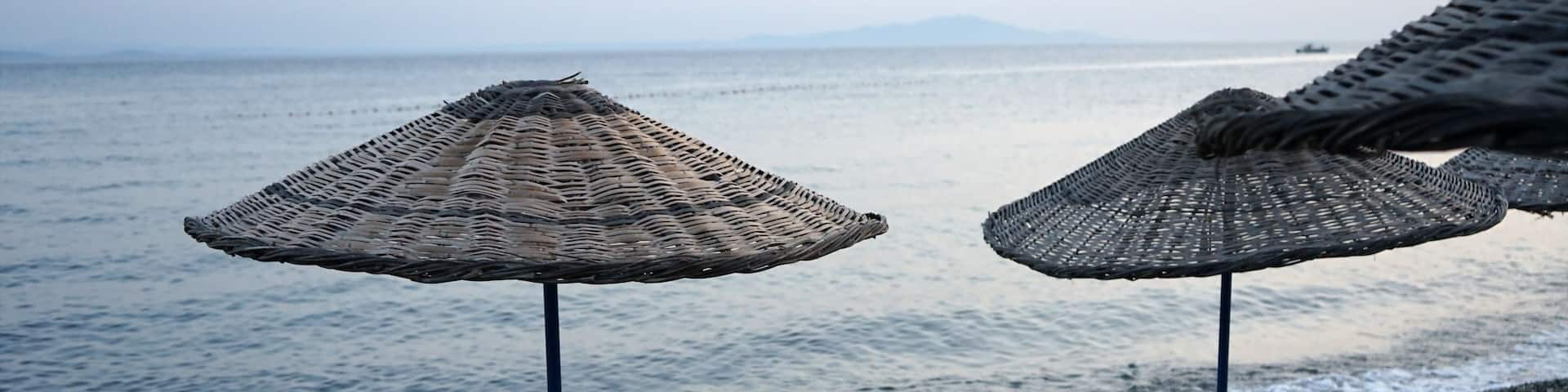 Blick vom Strand in Altinoluk mit Sonnenschirm aus Korbgeflecht im romantischen Licht der untergehenden Sonne auf den Golf von Edremit am Ägäischen Meer in der Provinz Balikesir in der Türkei