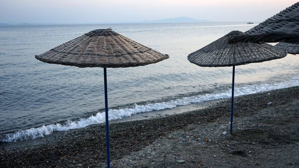 A beach with two woven umbrellas on poles, the sea, and distant mountains.