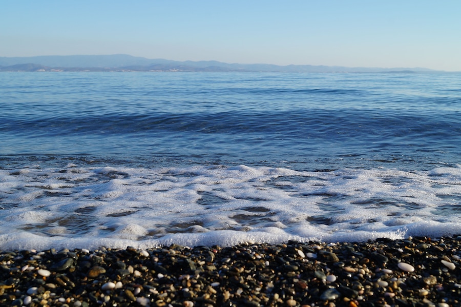 A pebbly beach with calm sea waves and distant mountains.