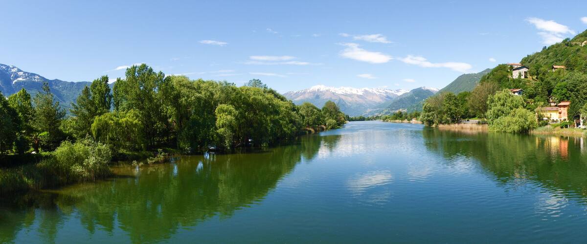 A river flanked by lush greenery and houses, with snow-capped mountains in the background.