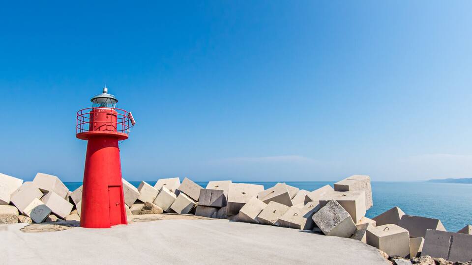 A red lighthouse with a metal railing, surrounded by concrete blocks, overlooking a calm sea.