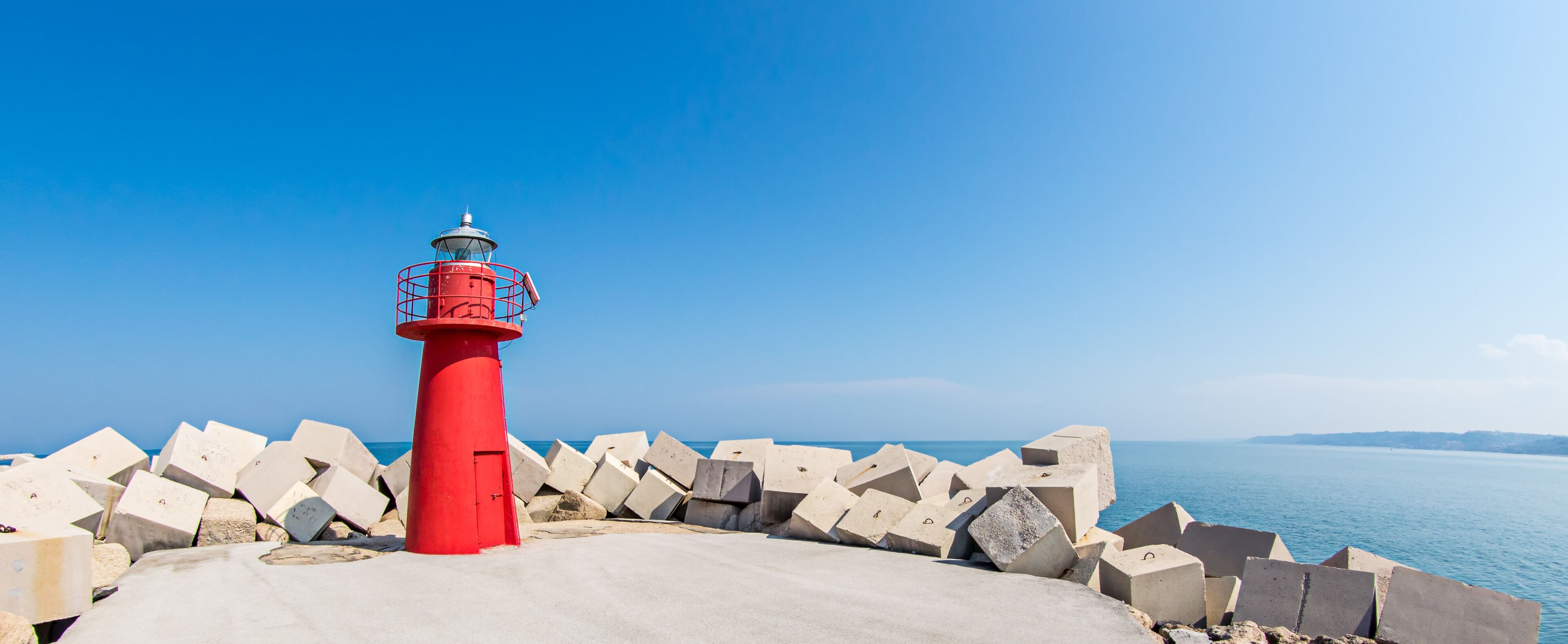 A red lighthouse with a metal railing, surrounded by concrete blocks, overlooking a calm sea.