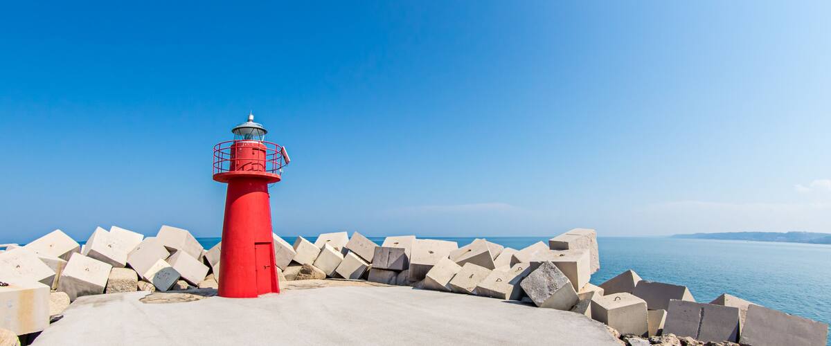 A red lighthouse with a metal railing, surrounded by concrete blocks, overlooking a calm sea.