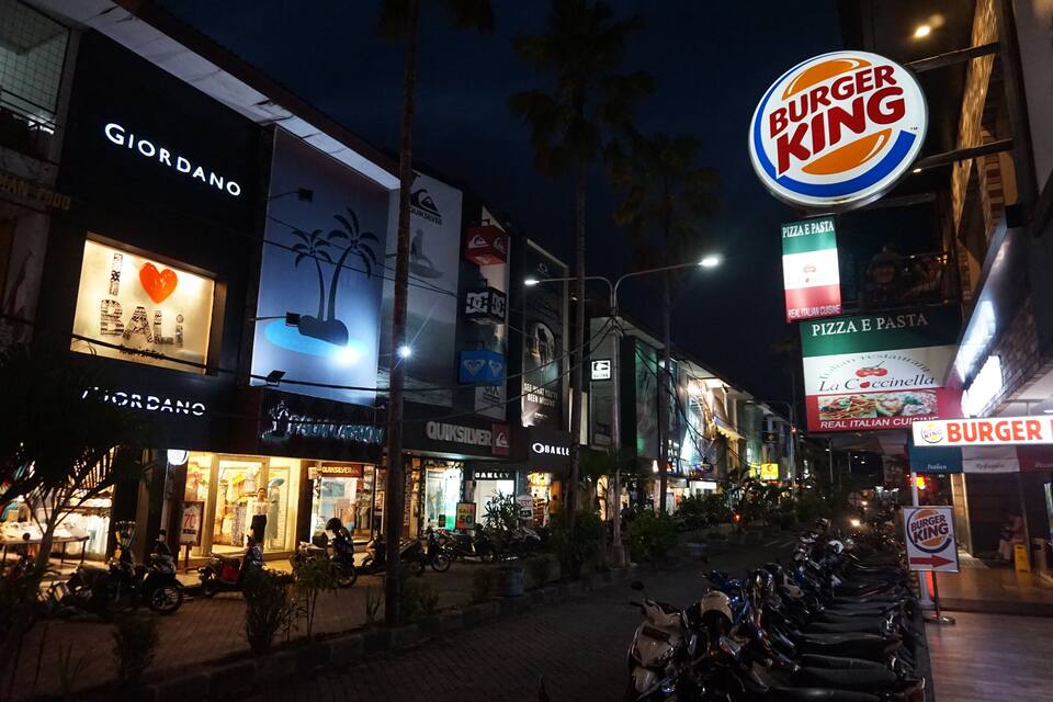 A street at night with various shops, including a Burger King and a pizza place.