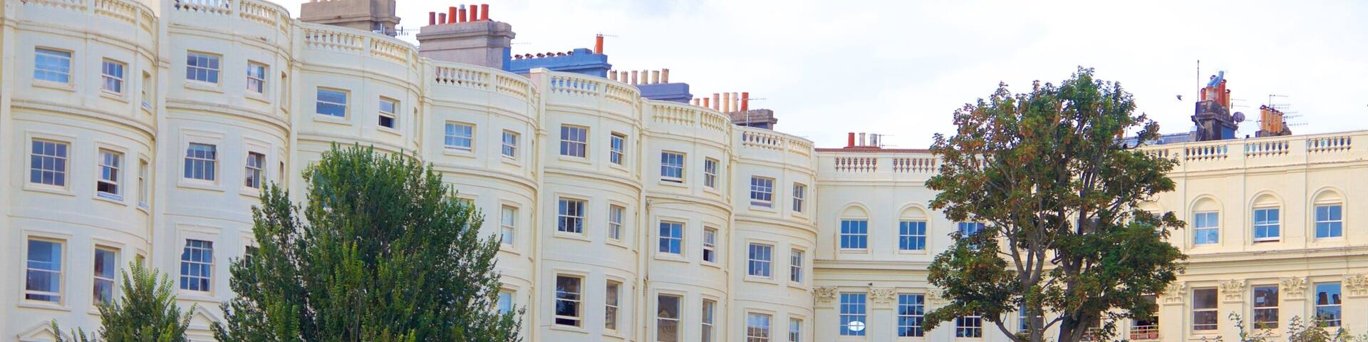 A row of cream-colored buildings with multiple windows and balconies, surrounded by lush greenery and trees.