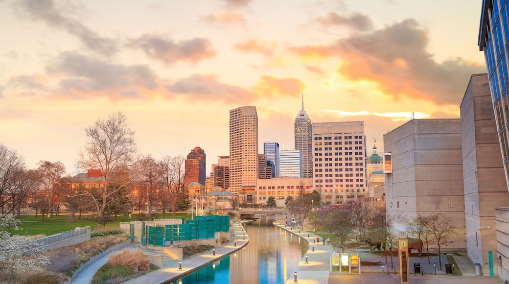 A cityscape with a river, modern buildings, and a park area during sunset.