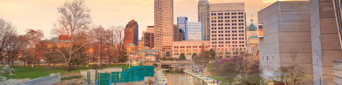 A cityscape with a river, modern buildings, and a park area during sunset.