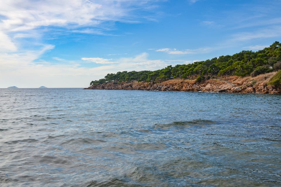 A coastal landscape with clear blue water, rocky cliffs, and dense green vegetation.