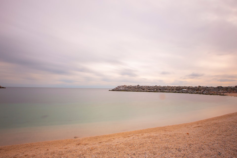 Een strand met kalm water, een kiezelstrand en een rotsachtige uitstulping in de verte.