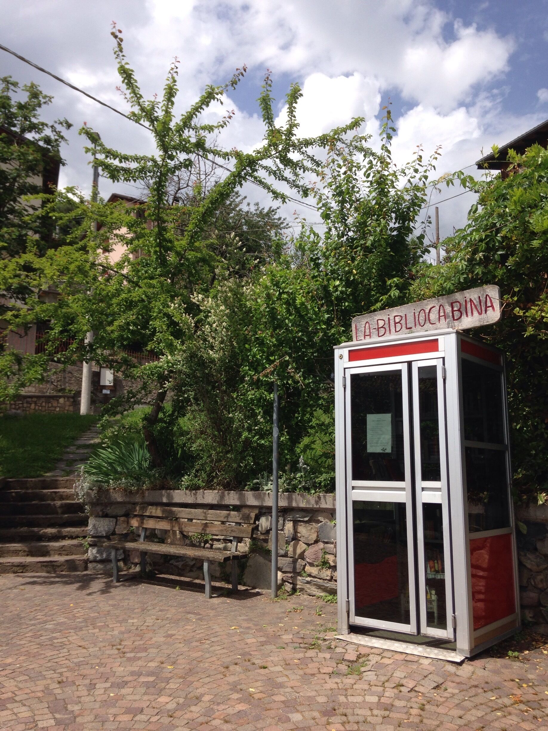 A public phone booth with a sign reading "La BiblioCA BINA" in a park with a stone wall, a bench, and trees.