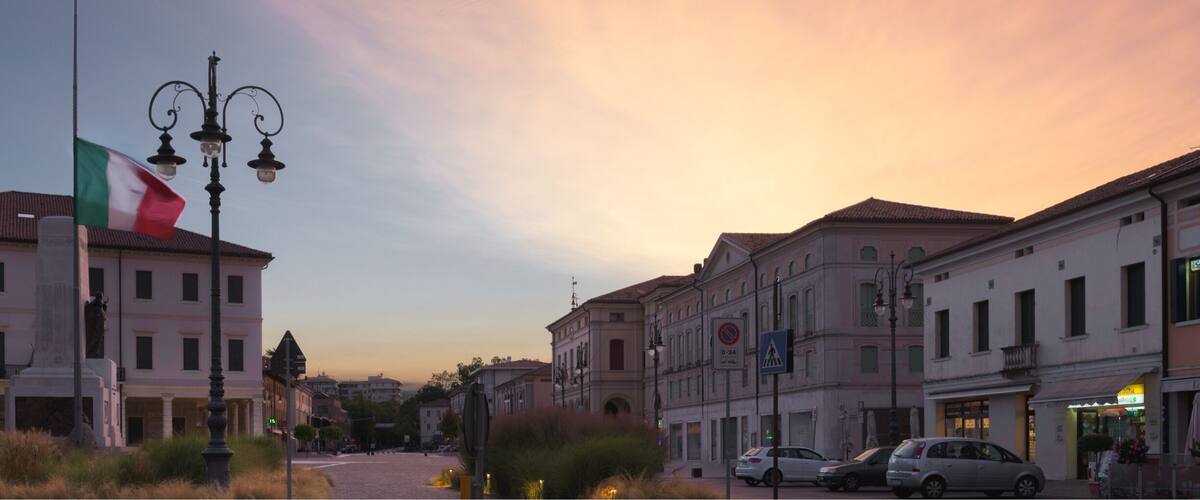 A sunset over a European town with historic buildings, a flag, and a statue.