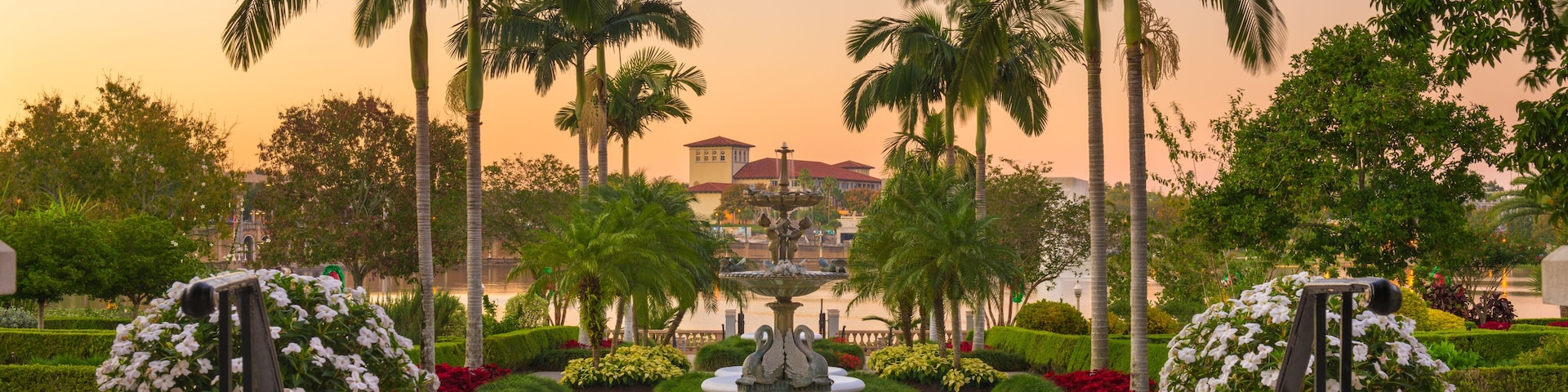 A circular fountain with a statue in the center, surrounded by palm trees and a building with a red roof in the background.