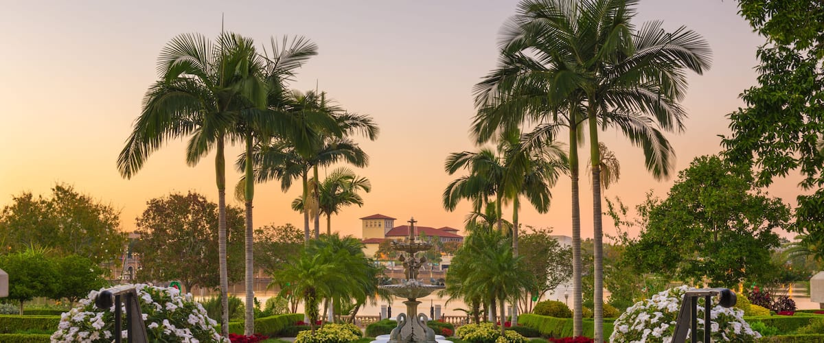 A circular fountain with a statue in the center, surrounded by palm trees and a building with a red roof in the background.
