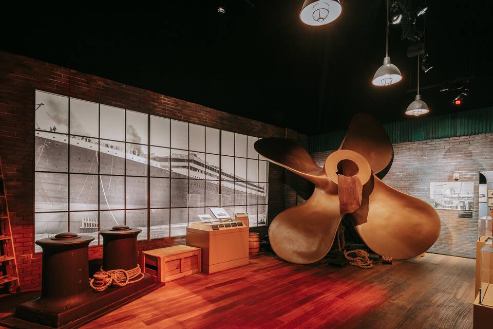 A museum exhibit with a large ship's anchor, wooden crates, and a ship's wheel.