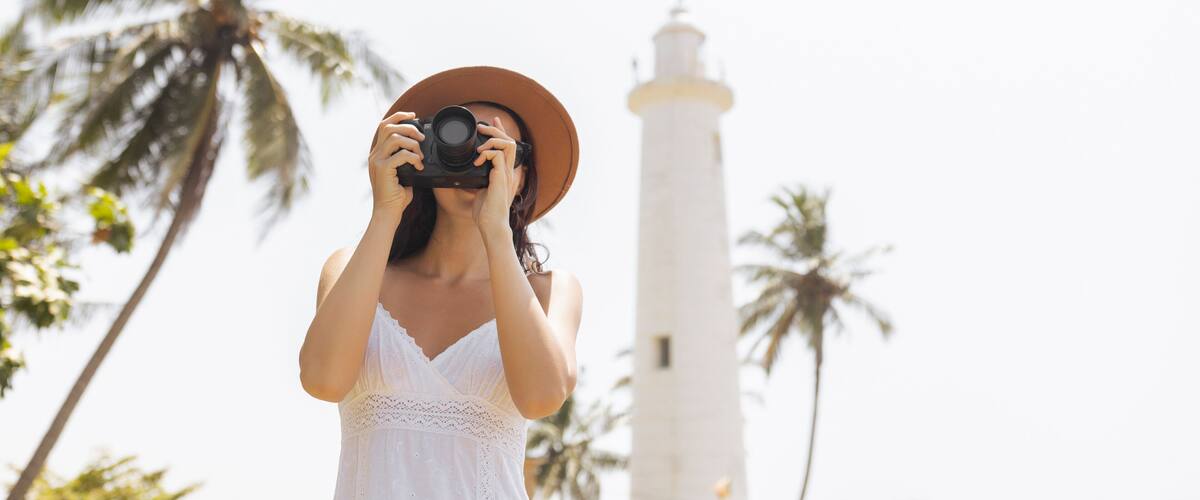 A person in a white dress and hat taking a photo with a camera, palm trees, and a lighthouse in the background.