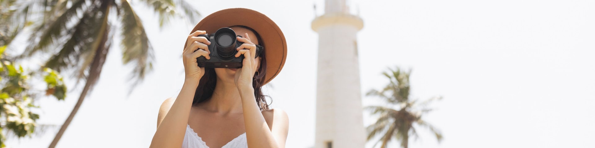 A person in a white dress and hat taking a photo with a camera, palm trees, and a lighthouse in the background.
