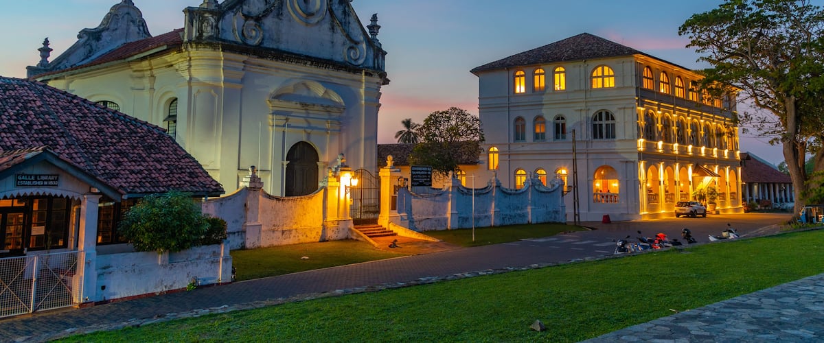 Un edificio histórico con arquitectura ornamentada y un jardín bien cuidado al anochecer.