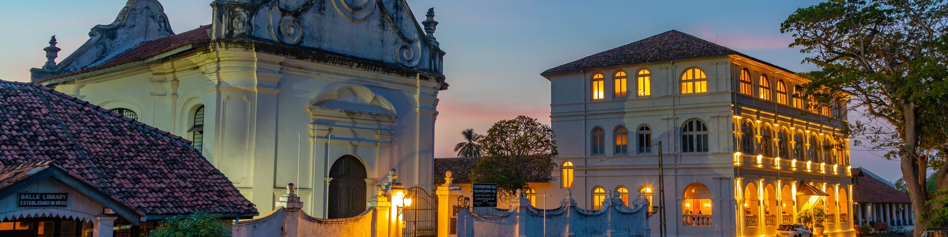 Un edificio histórico con arquitectura ornamentada y un jardín bien cuidado al anochecer.