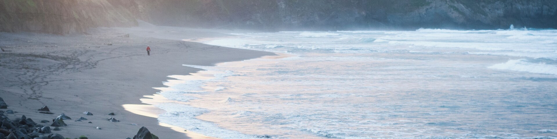 A rocky beach with waves crashing onto the shore and a person walking along the water's edge.