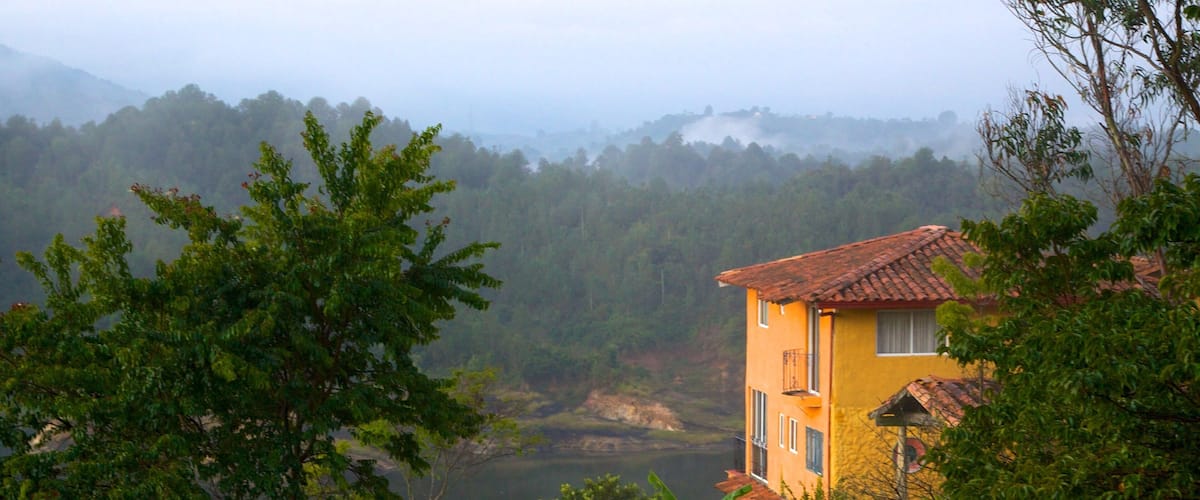 A yellow house with a red roof, surrounded by lush greenery and a lake.