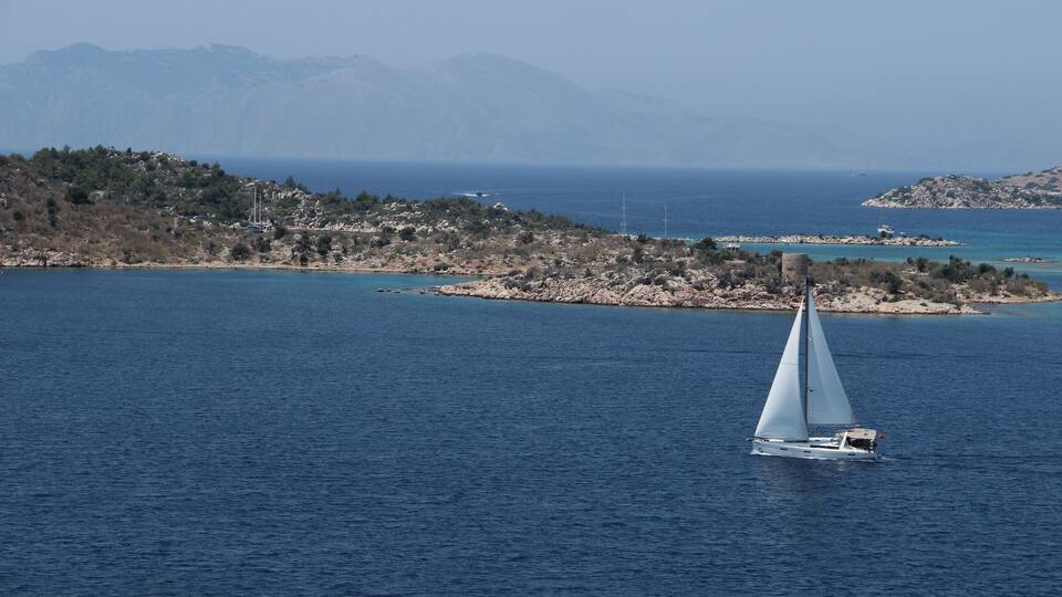 A sailboat on a calm sea with a mountainous coastline in the background.