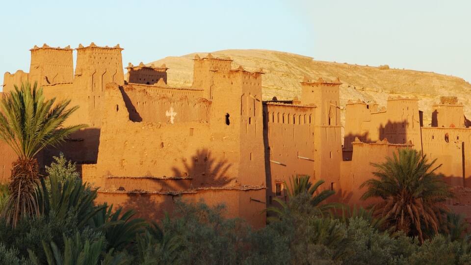 A large, ancient fort with multiple towers and a hill in the background. Palm trees are in the foreground.
