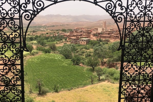 A view through a wrought iron gate of a rural landscape with green fields, scattered trees, and distant buildings.