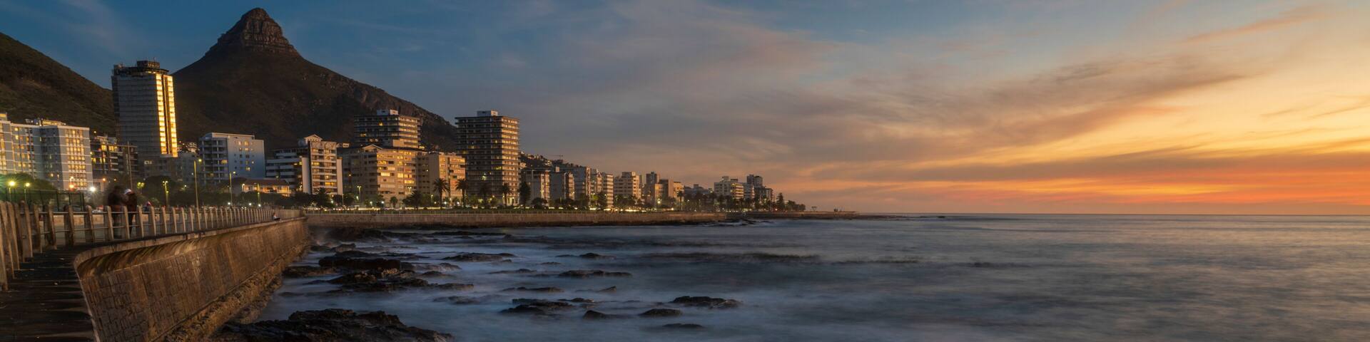 A coastal cityscape at sunset with a prominent mountain in the background and a rocky shoreline.