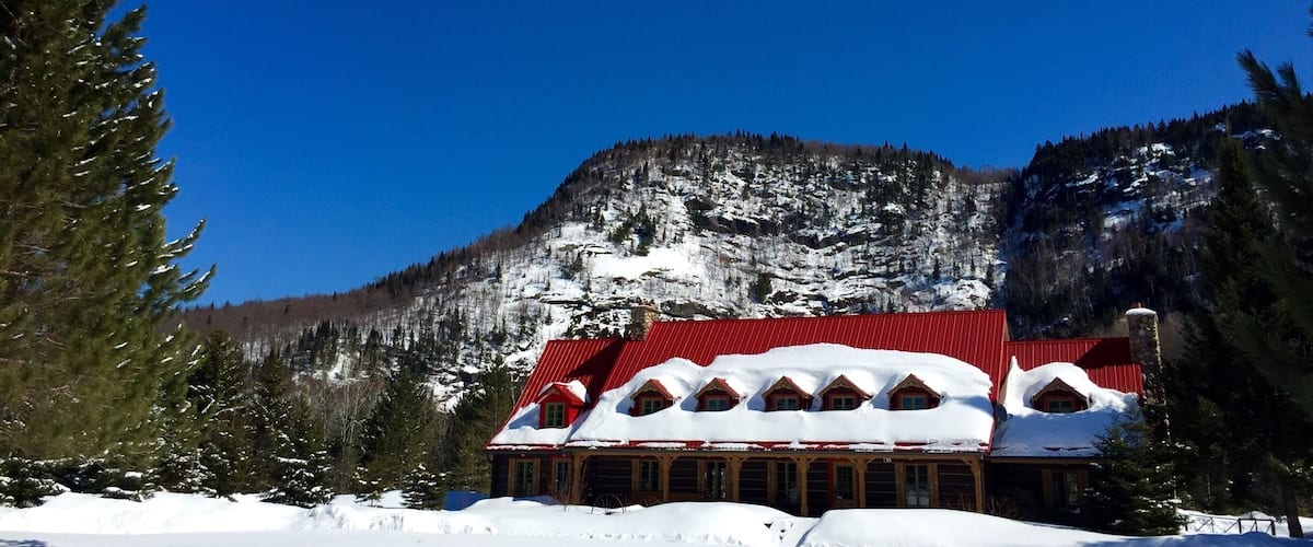 A snow-covered lodge with a red roof, surrounded by snow-laden trees and a mountain backdrop.