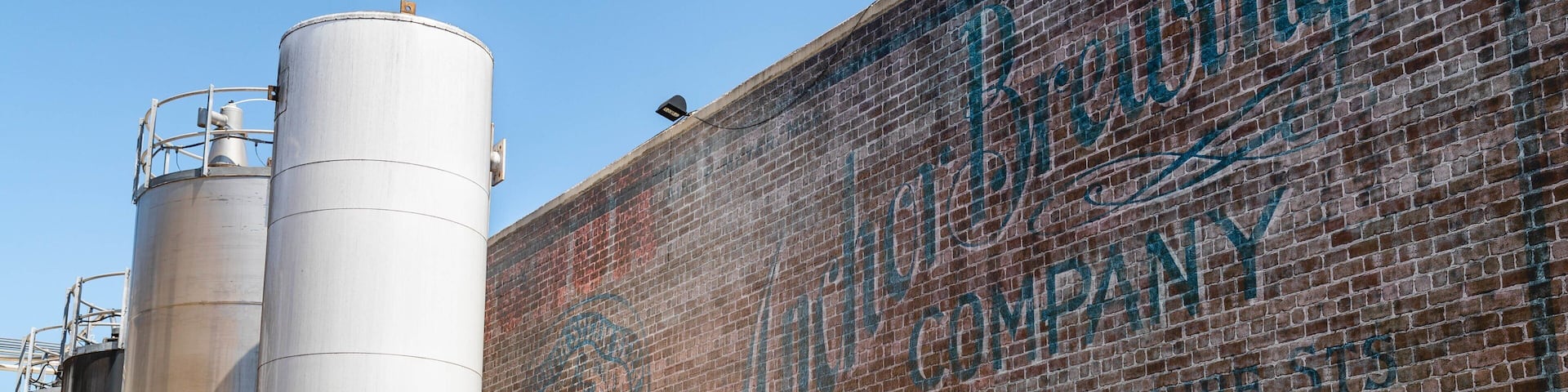 A brick wall with a mural advertising the Brewing Company, large cylindrical storage tanks, and a clear blue sky.