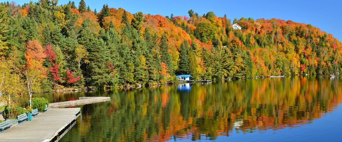 A calm lake with a wooden dock and a house on a hill surrounded by autumn-colored trees.