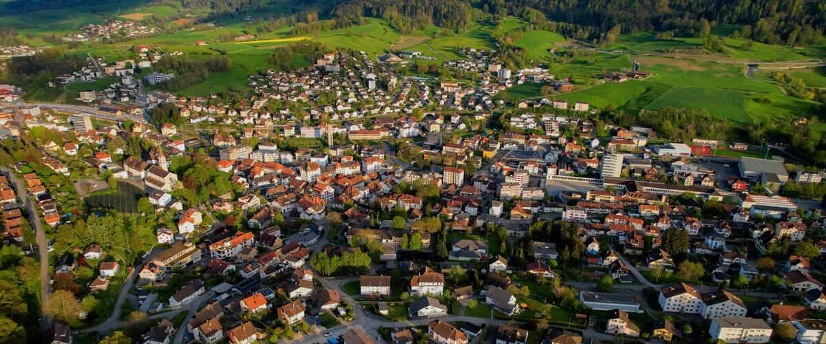 An aerial view of a densely populated town with a mix of residential houses and commercial buildings, surrounded by green fields and hills.